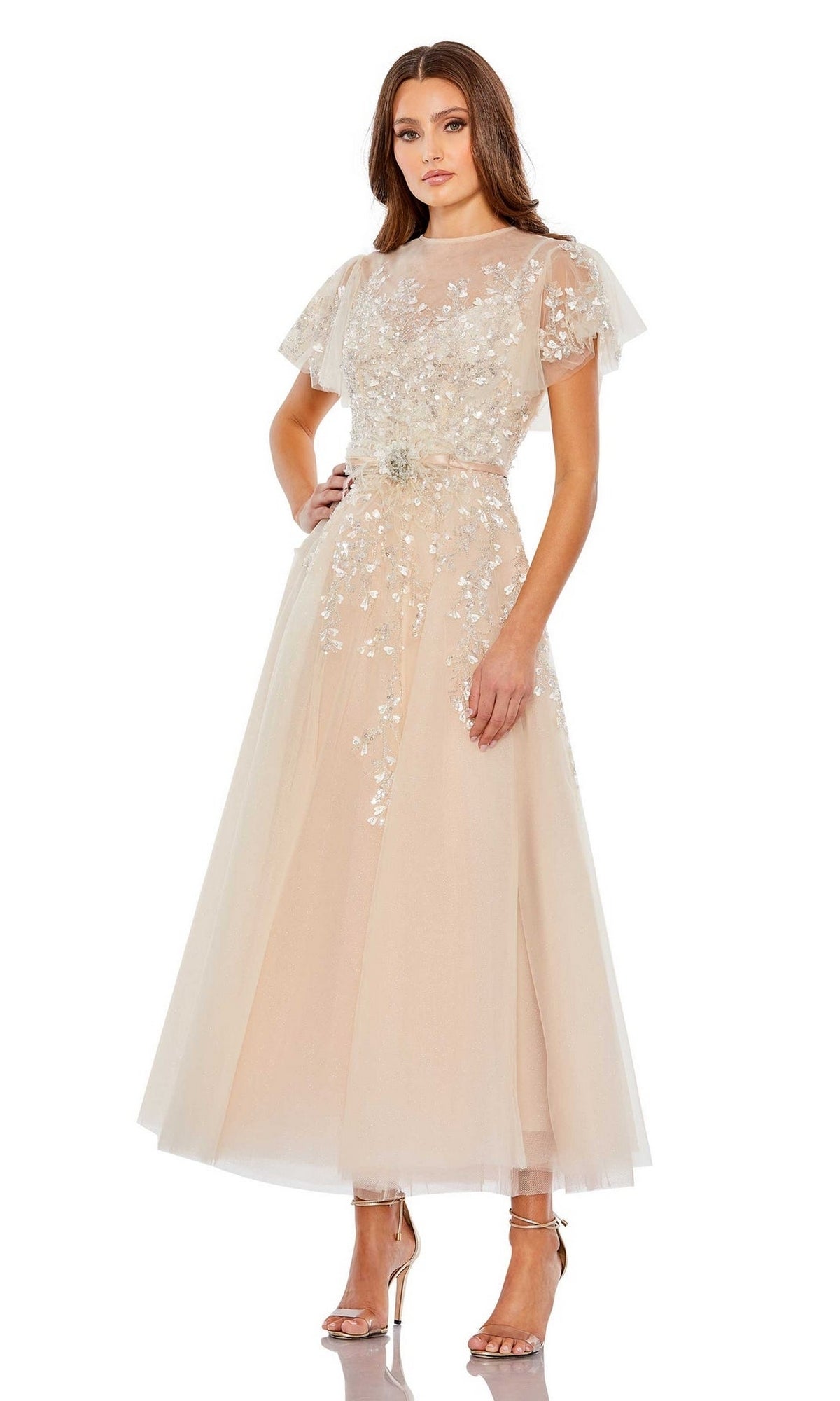 A woman wears the Long Formal Dress 20362 by Mac Duggal, a light beige tea-length A-line gown with floral embroidery, flutter sleeves, and high-heeled sandals.